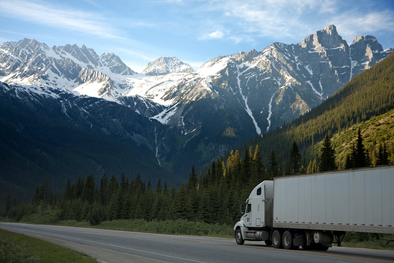 about-01 A semi-truck travels along a highway with snow-capped mountains in the background.